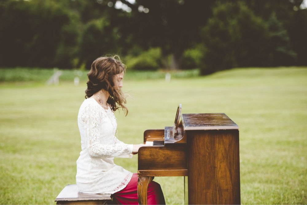 Brown haired woman in a white blouse with red pants playing piano in a field
