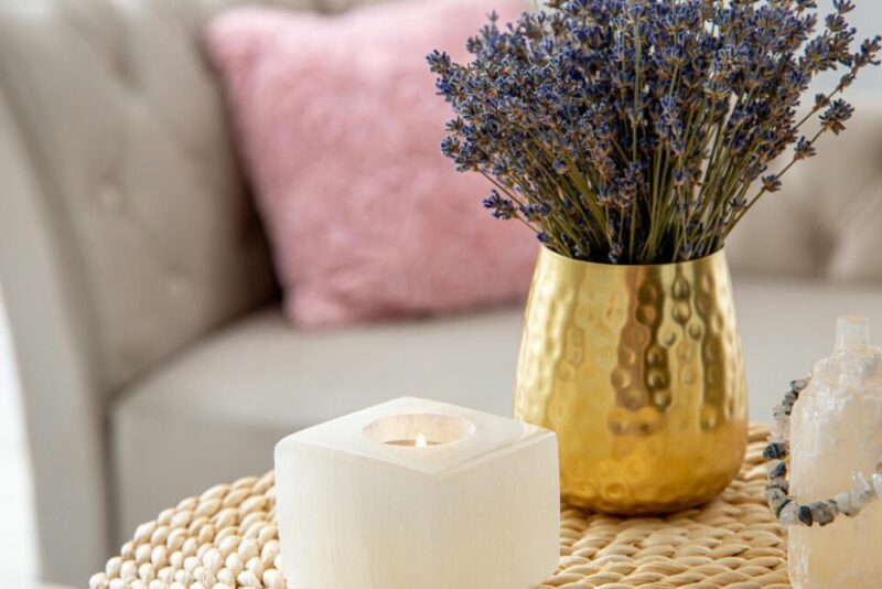 White lit candle and gold vase with lavender on a wicker table. A grey couch with a pink pillow is in the background